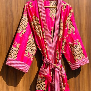 May include: A vibrant pink robe with a tiger print. The robe features a striped pattern on the collar, cuffs, and belt. The tiger print is in shades of tan and black. The robe is hanging on a wooden hanger.