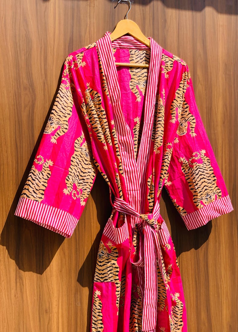 May include: A vibrant pink robe with a tiger print. The robe features a striped pattern on the collar, cuffs, and belt. The tiger print is in shades of tan and black. The robe is hanging on a wooden hanger.