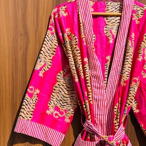 May include: A vibrant pink robe with a tiger print and striped accents. The robe features a repeating pattern of tigers in shades of tan and black. The trim and stripes are white and red. The robe is tied at the waist with a matching belt.