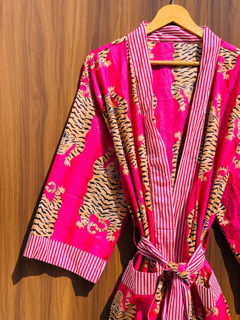 May include: A vibrant pink robe with a tiger print and striped accents. The robe features a repeating pattern of tigers in shades of tan and black. The trim and stripes are white and red. The robe is tied at the waist with a matching belt.