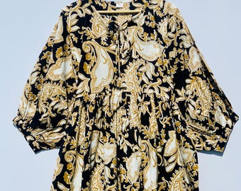 May include: A black dress with a white and gold paisley pattern. The dress has long sleeves, a gathered waist, and a tie-neck detail. The fabric appears to be lightweight, suitable for warmer weather.