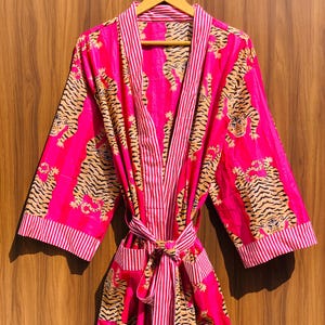 May include: A vibrant pink robe with a tiger print and striped accents. The robe features a tie closure, pockets, and a striped trim. The tiger pattern is in shades of brown and black. The robe is hanging on a wooden hanger.
