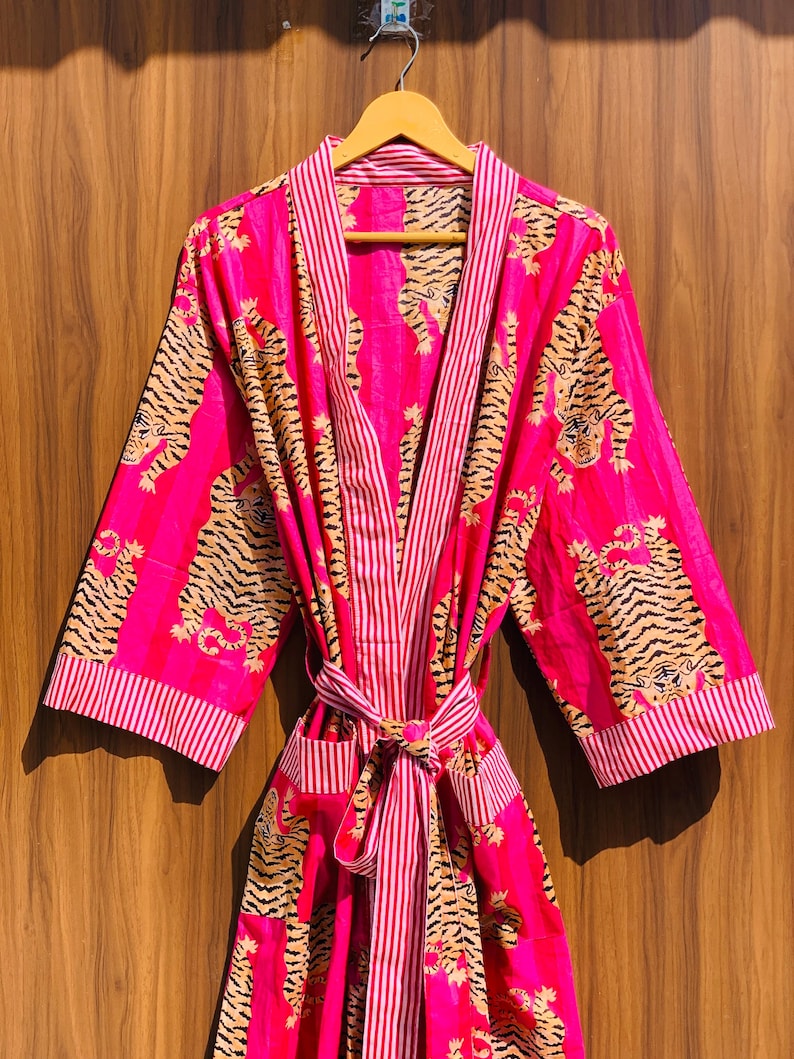 May include: A vibrant pink robe with a tiger print and striped accents. The robe features a tie closure, pockets, and a striped trim. The tiger pattern is in shades of brown and black. The robe is hanging on a wooden hanger.