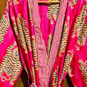 May include: A vibrant pink robe with a tiger print and red and white striped accents. The robe features a tie belt and is displayed on a wooden hanger. The tiger print includes black stripes and tan fur.