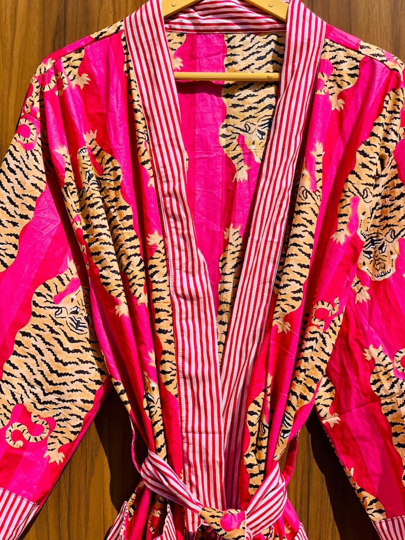 May include: A vibrant pink robe with a tiger print and red and white striped accents. The robe features a tie belt and is displayed on a wooden hanger. The tiger print includes black stripes and tan fur.
