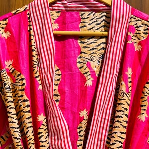 May include: A vibrant pink robe with a tiger print. The robe features a bold tiger pattern in black and tan against a pink background. The collar and trim have red and white stripes. The robe is hanging on a wooden hanger.