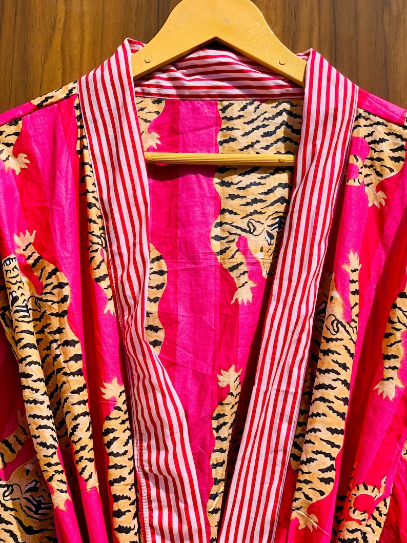 May include: A vibrant pink robe with a tiger print. The robe features a bold tiger pattern in black and tan against a pink background. The collar and trim have red and white stripes. The robe is hanging on a wooden hanger.