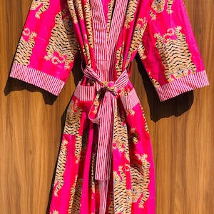 May include: A vibrant pink robe with a tiger print. The robe features a striped pattern on the lapels, cuffs, and belt. The tiger print is in shades of tan and black. The robe is tied at the waist with a matching belt.