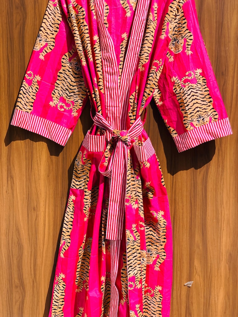 May include: A vibrant pink robe with a tiger print. The robe features a striped pattern on the lapels, cuffs, and belt. The tiger print is in shades of tan and black. The robe is tied at the waist with a matching belt.