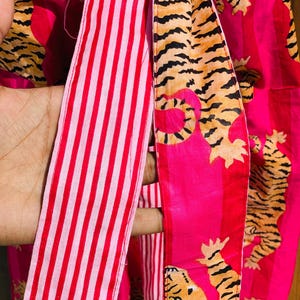 May include: Close-up of a vibrant pink fabric with a tiger stripe pattern and red and white striped ribbon. The tiger stripes are in shades of orange and black. The fabric is likely used for clothing or accessories.