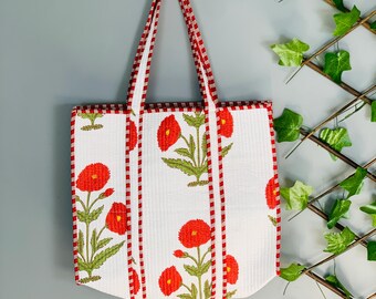 May include: White tote bag with a red and green floral pattern. The bag features red trim and handles. The floral design includes red flowers and green leaves. The bag is hanging on a wooden hook against a gray wall.