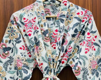May include: A cream-colored robe with a floral pattern in red, gray, and gold hangs on a wooden hanger. The robe features a tie closure at the waist and long sleeves. The floral design includes large, stylized flowers and leaves.