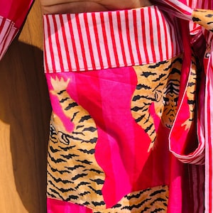 May include: A vibrant pink robe with a tiger print design. The robe features a striped collar and tie, and the tiger print is in shades of tan and black. The robe is made of a lightweight fabric.