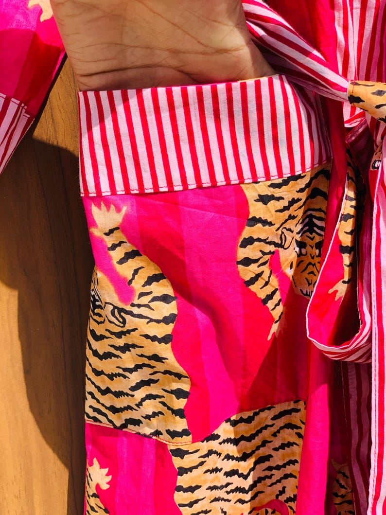 May include: A vibrant pink robe with a tiger print design. The robe features a striped collar and tie, and the tiger print is in shades of tan and black. The robe is made of a lightweight fabric.