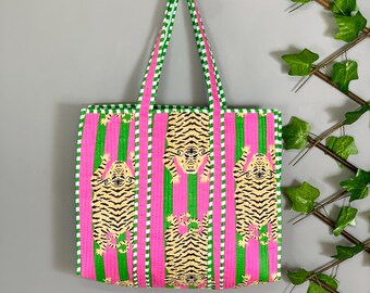 May include: A vibrant tote bag with a tiger print. The bag features vertical stripes in pink and green, with a repeating pattern of tigers. The handles and trim are green and white. The bag is hanging on a wooden hook.