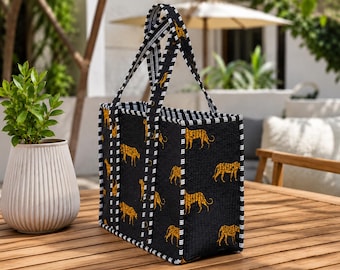 May include: A rectangular tote bag featuring a black fabric base with a repeating pattern of yellow and orange cheetahs. The bag has two upright handles and edges trimmed with a black and white striped fabric pattern throughout the design.
