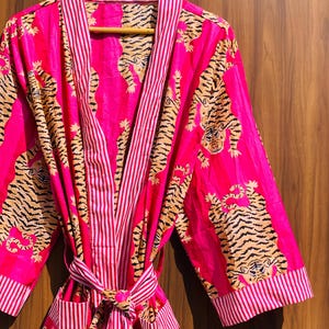 May include: A vibrant pink robe with a tiger print and striped accents. The robe features a repeating pattern of tigers in shades of brown and black. The trim and belt are striped in pink and white. The robe is hanging on a wooden hanger.
