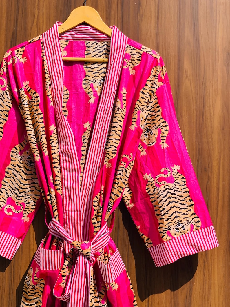 May include: A vibrant pink robe with a tiger print and striped accents. The robe features a repeating pattern of tigers in shades of brown and black. The trim and belt are striped in pink and white. The robe is hanging on a wooden hanger.