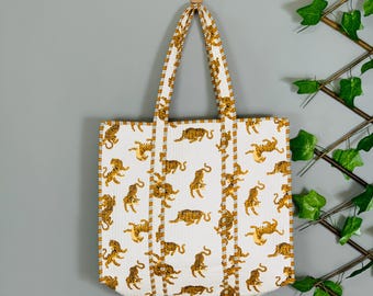 May include: A white quilted tote bag featuring a repeating pattern of orange and black tigers. The bag has two long shoulder straps with a white and mustard yellow striped trim that matches the edges of the rectangular bag body.