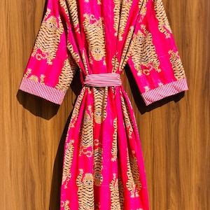 May include: A vibrant pink robe with a repeating tiger print in shades of tan and black. The robe features a matching belt and striped trim on the collar and sleeves. The garment is hanging on a wooden hanger.