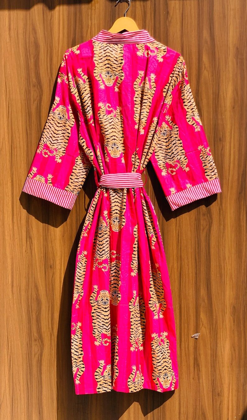 May include: A vibrant pink robe with a repeating tiger print in shades of tan and black. The robe features a matching belt and striped trim on the collar and sleeves. The garment is hanging on a wooden hanger.