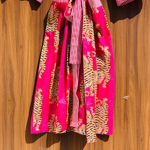 May include: A vibrant pink robe with a tiger print and striped accents. The robe features a tie closure and wide sleeves. The tiger pattern is in shades of black and gold. The striped details are in pink and white.