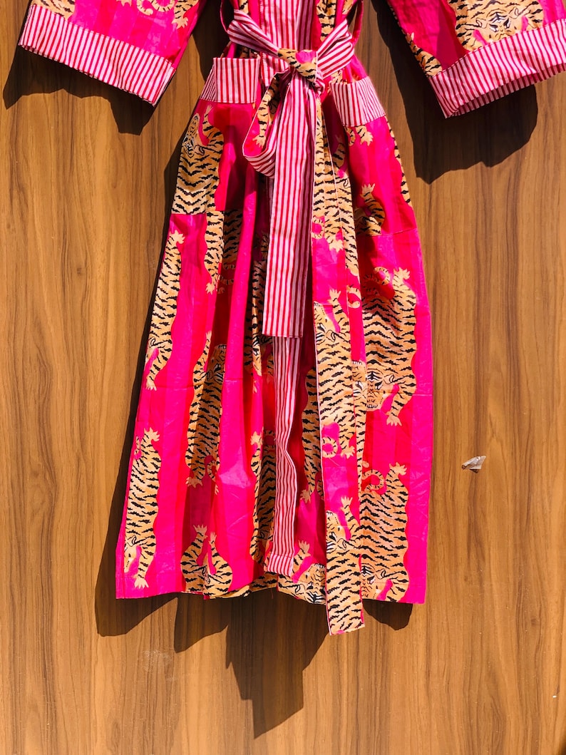 May include: A vibrant pink robe with a tiger print and striped accents. The robe features a tie closure and wide sleeves. The tiger pattern is in shades of black and gold. The striped details are in pink and white.
