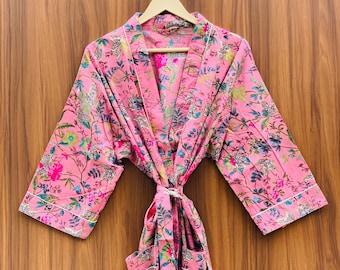 May include: A pink floral patterned robe hanging on a wooden hanger. The robe features a vibrant design with flowers and leaves in shades of pink, green, and blue. White trim accents the edges and sleeves. The robe is tied at the waist with a matching fabric belt.