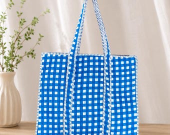 May include: A rectangular tote bag featuring a vibrant blue and white gingham check pattern. The bag is constructed with quilted fabric and includes two matching blue and white checkered handles that extend from the base to the top opening.