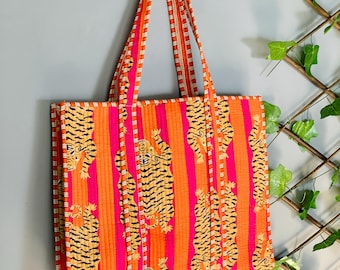 May include: A vibrant tote bag with a tiger print. The bag features vertical stripes in shades of orange and pink, with tiger illustrations. The handles are orange and white striped. The bag is hanging against a gray wall.