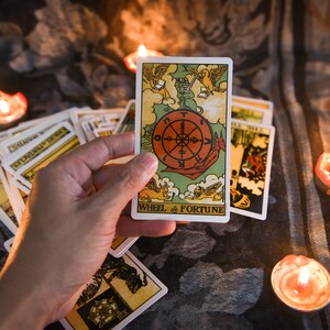 May include: A tarot card reading with the Wheel of Fortune card being held up. The card is surrounded by other tarot cards and lit candles.