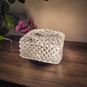 May include: A clear, square glass ashtray with a diamond-cut design. The ashtray is on a black base and is lit, reflecting light. The background features a wooden surface, a vase with greenery, and a purple orchid.