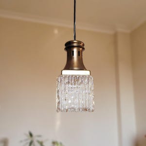 May include: A brass and glass pendant light with a square, clear glass shade featuring vertical lines. The light is hanging from a black wire.