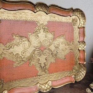 May include: A vintage, decorative tray with a peach-colored base and gold-leaf accents. The tray features a raised, ornate design in the center and a scalloped edge.