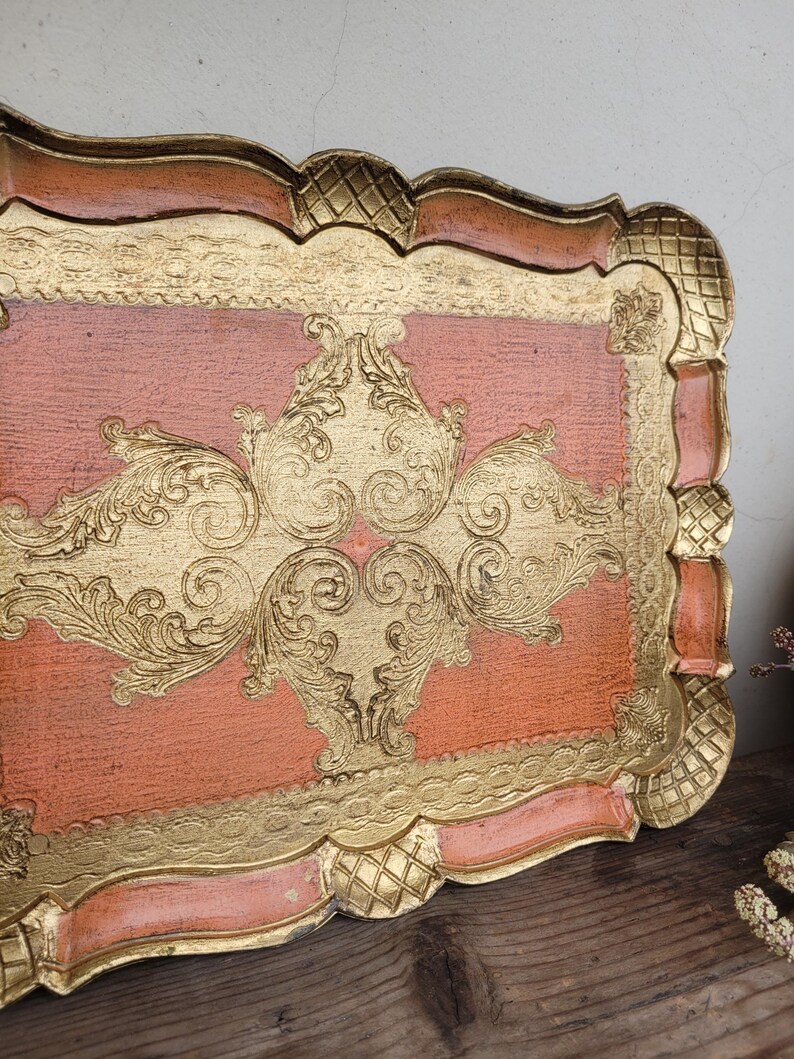 May include: A vintage, decorative tray with a peach-colored base and gold-leaf accents. The tray features a raised, ornate design in the center and a scalloped edge.