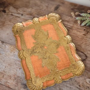 May include: A decorative tray with a peach-colored base and gold-colored intricate floral design. The tray has a scalloped edge.