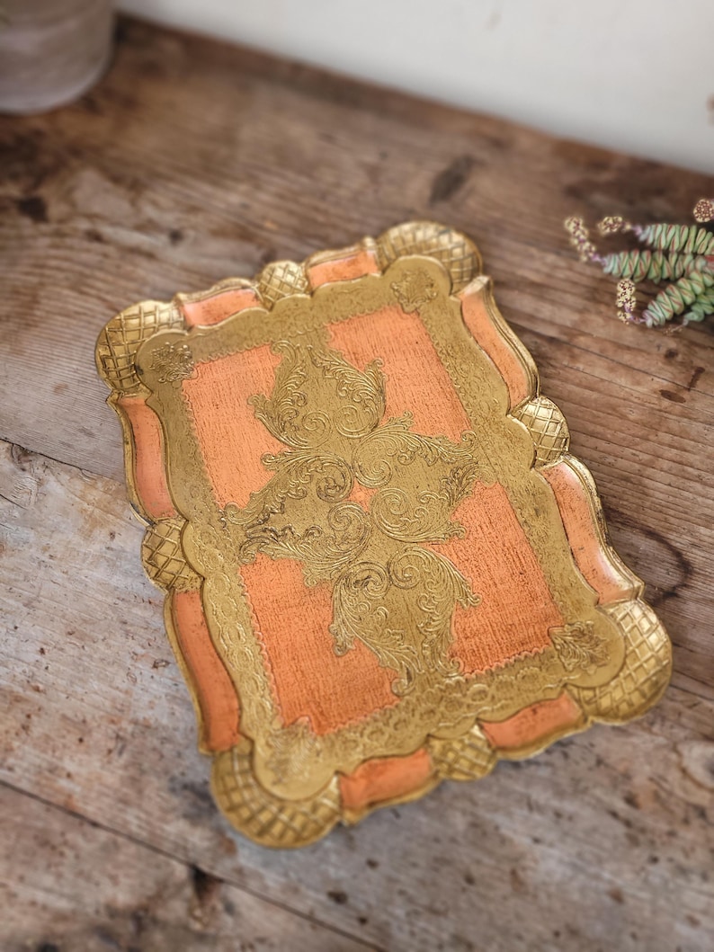 May include: A decorative tray with a peach-colored base and gold-colored intricate floral design. The tray has a scalloped edge.