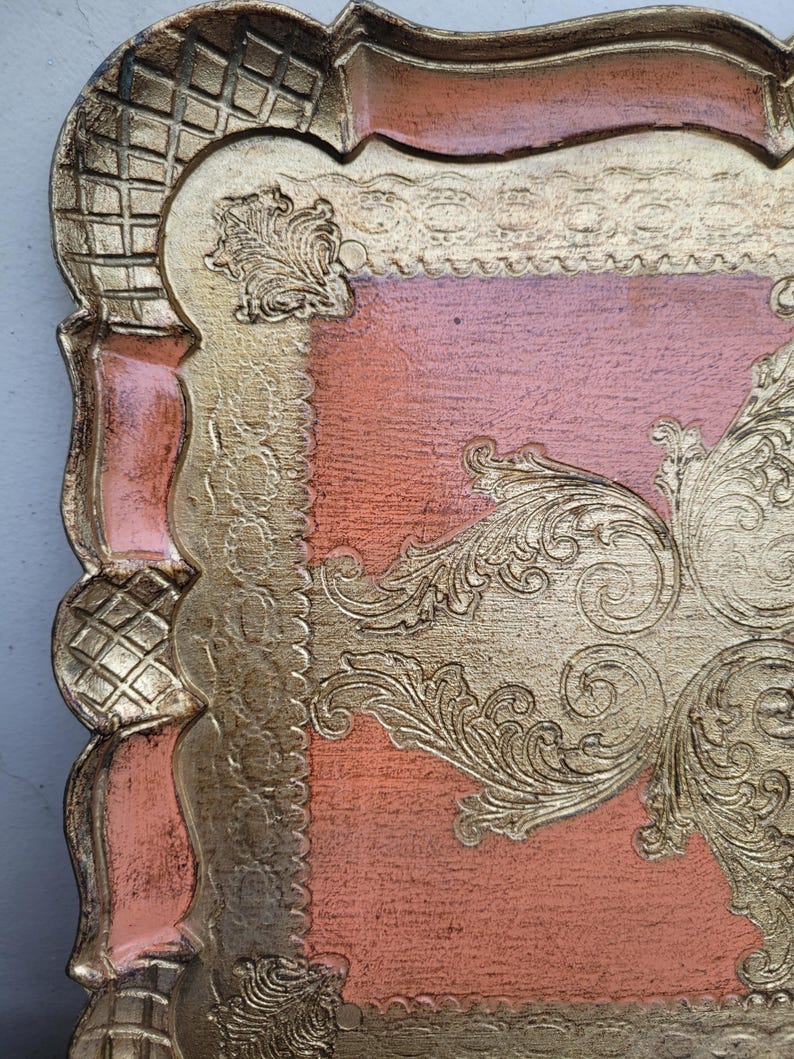 May include: A square, decorative tray with a raised, ornate design. The tray is made of wood and has a pink and gold finish.