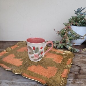 May include: A pink and white ceramic mug with a floral design and a gold rim sits on a gold and orange tray with a decorative border.