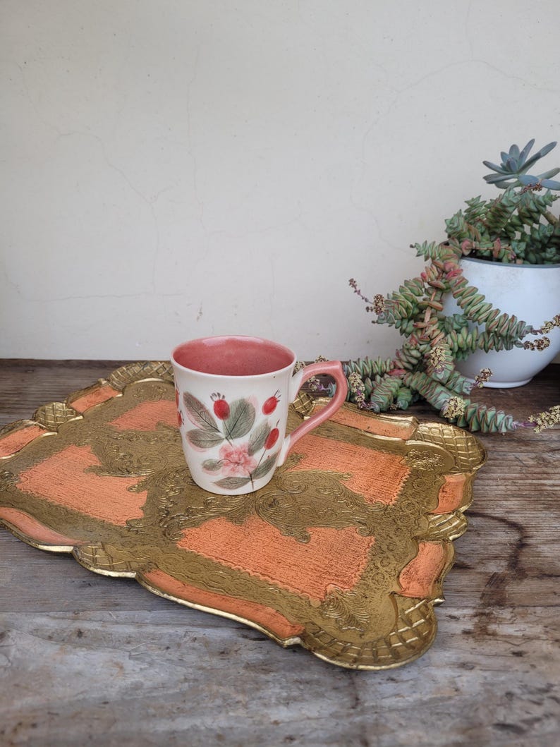 May include: A pink and white ceramic mug with a floral design and a gold rim sits on a gold and orange tray with a decorative border.