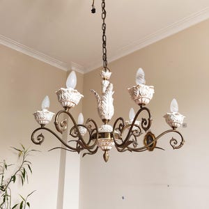 May include: An ornate chandelier with six lights. The fixture features white, decorative candle holders and a central column with a floral design. The metal frame has a bronze finish. The chandelier hangs from a chain.