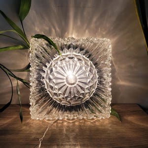 May include: A clear glass square ceiling light fitting with a diamond pattern. The light is on and casting a warm glow.