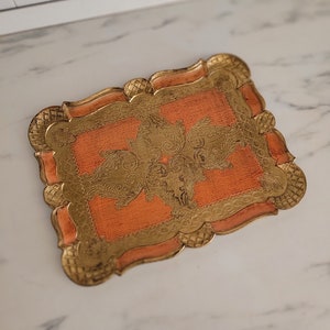 May include: A decorative tray with a peach-colored base and gold-colored intricate design. The tray has a scalloped edge.