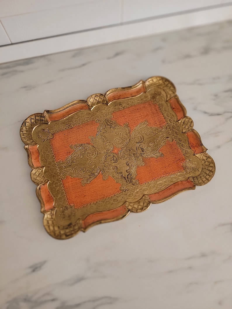 May include: A decorative tray with a peach-colored base and gold-colored intricate design. The tray has a scalloped edge.