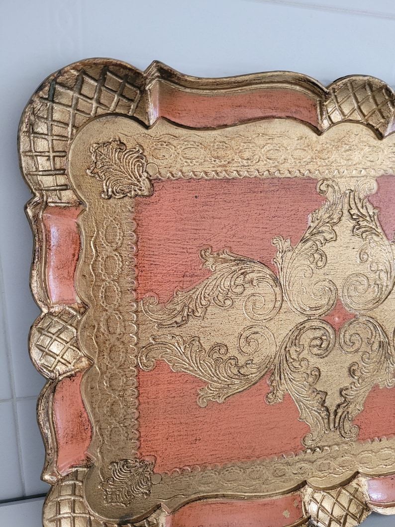 May include: A vintage tray with a pink painted surface and gold leaf accents. The tray has a decorative, ornate design with a scalloped edge.