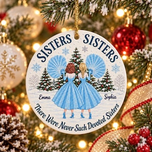 Op de afbeelding: Een ronde witte keramische hanger met twee figuren in blauwe jurken die blauwe waaiers vasthouden voor een kerstboom. De tekst luidt SISTERS SISTERS, Emma, Sophia en There Were Never Such Devoted Sisters, met blauwe sneeuwvlokken.