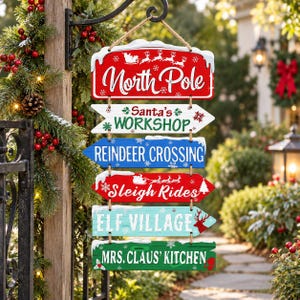 May include: A hanging Christmas sign featuring six stacked wooden directional arrows with snow-dusted edges. The signs read North Pole, Santa's Workshop, Reindeer Crossing, Sleigh Rides, Elf Village, and Mrs. Claus' Kitchen in red, white, blue, and green.