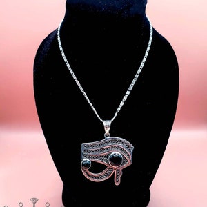 May include: Silver chain necklace with a silver pendant featuring a black eye of Horus design. The pendant has intricate filigree details and two black gemstones.