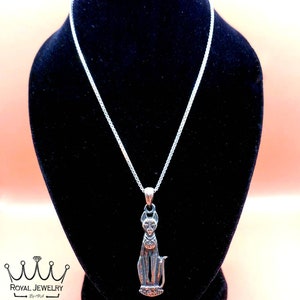 May include: Silver chain necklace with a silver pendant of a sitting cat. The pendant has intricate details and a textured surface. The necklace is displayed on a black velvet stand.