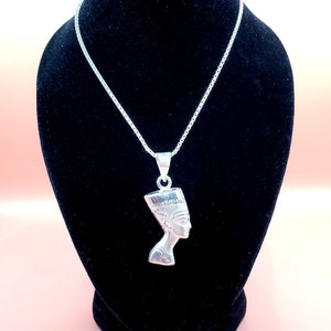 May include: A silver pendant necklace featuring a detailed profile of Nefertiti. The pendant is suspended from a delicate silver chain. The necklace is displayed on a black velvet bust against a pink background. The Royal Jewelry logo is visible.